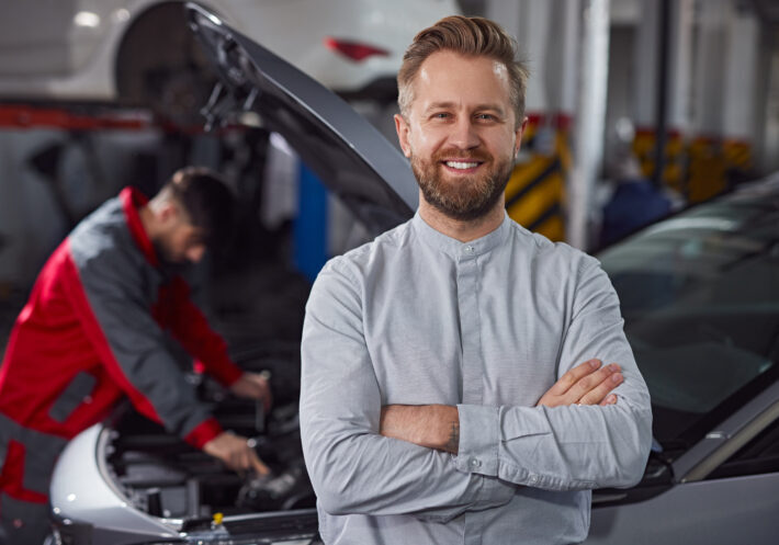 Positive bearded man with crossed arms smiling and looking at camera while standing near technician repairing vehicle in garage