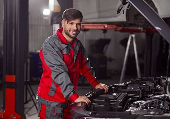 Side view of professional repairman in workwear looking at camera and smiling friendly while standing near car with open hood during diagnostic service in workshop